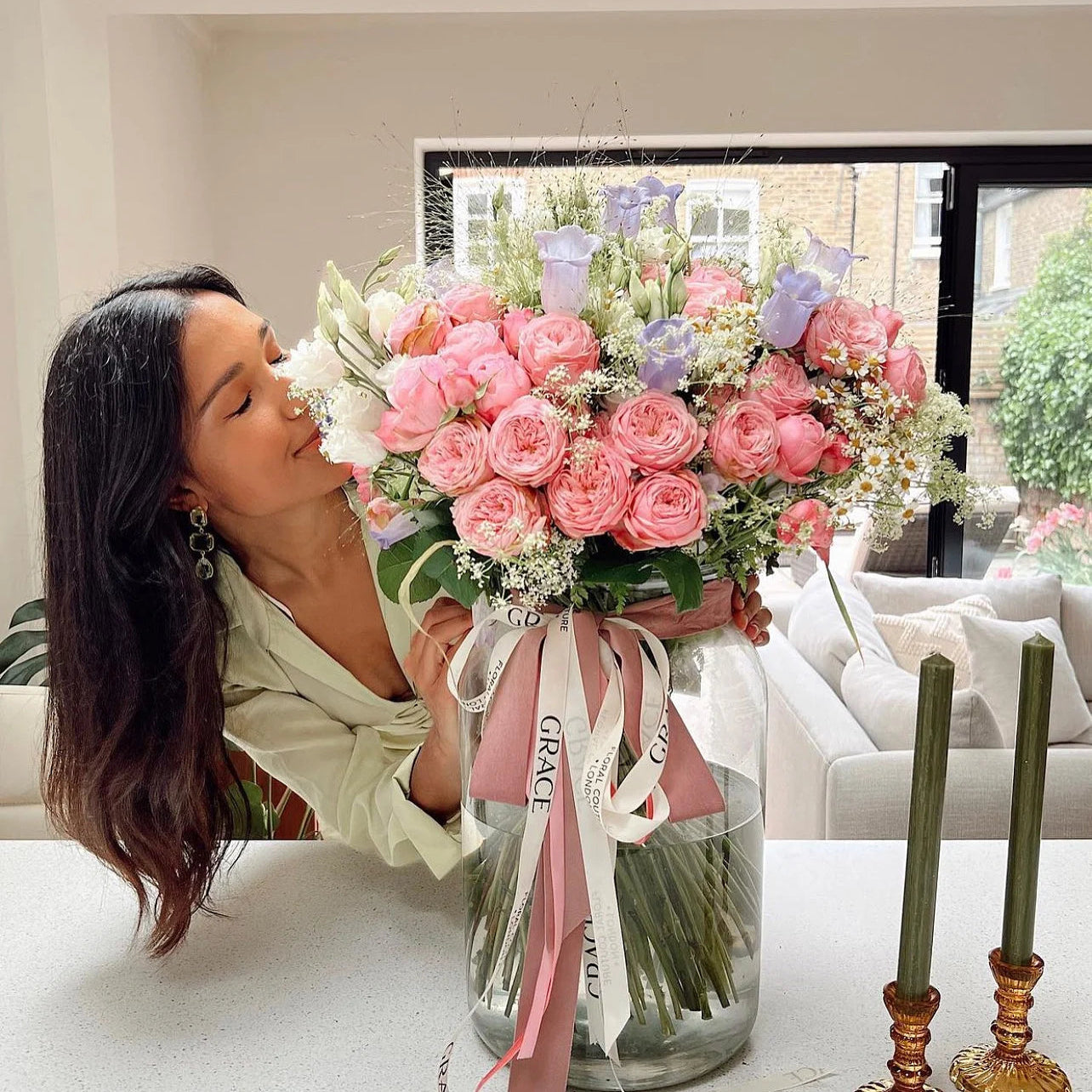 Woman kissing a large bouquet of pink roses with a 'Grace' ribbon in a living room.