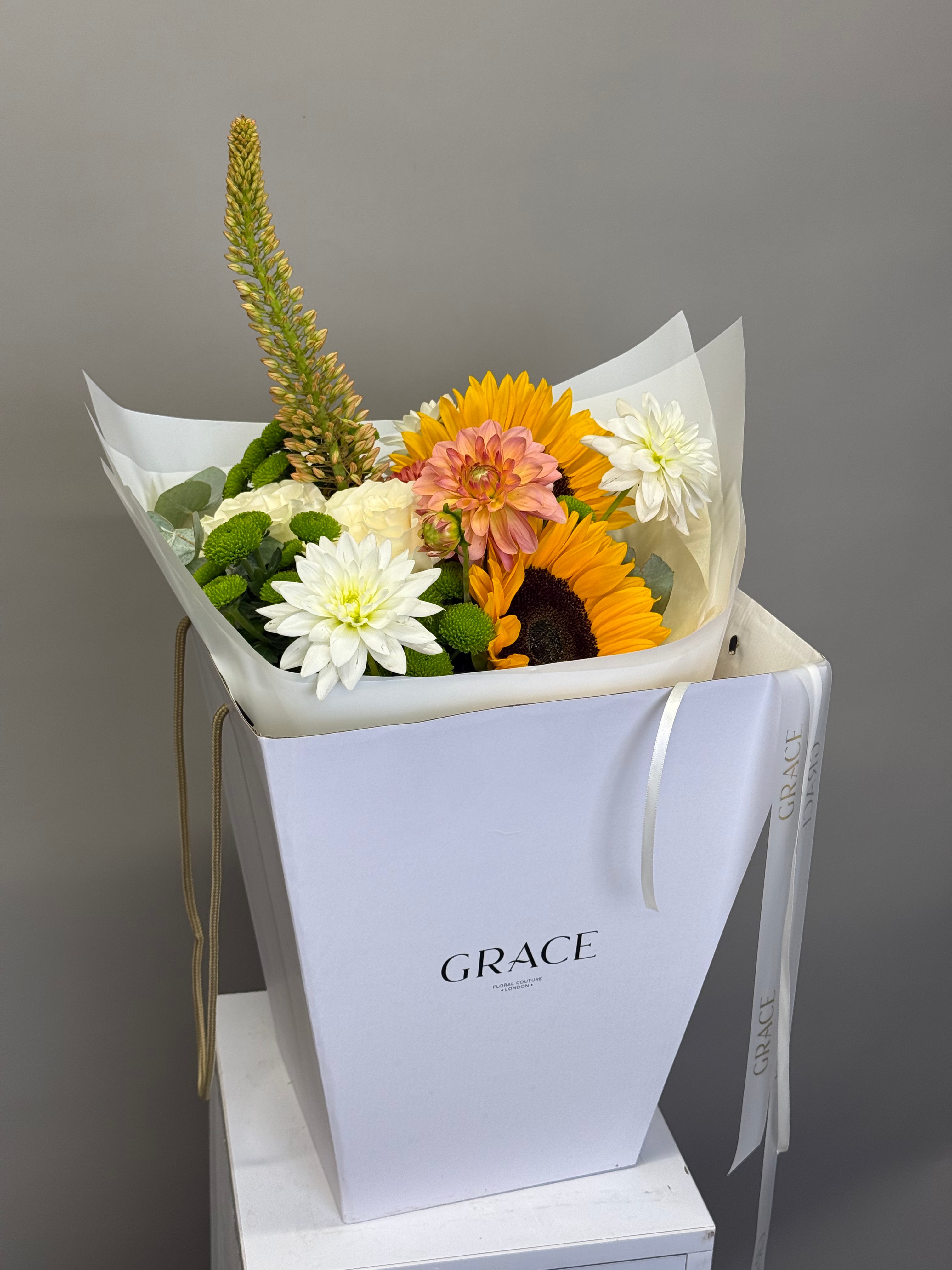 A floral bouquet in a white gift box with the brand name GRACE printed on the front. The bouquet features white dahlia-like flowers, a bright yellow sunflower, a peach-pink dahlia, and green foliage, wrapped in white tissue paper.
