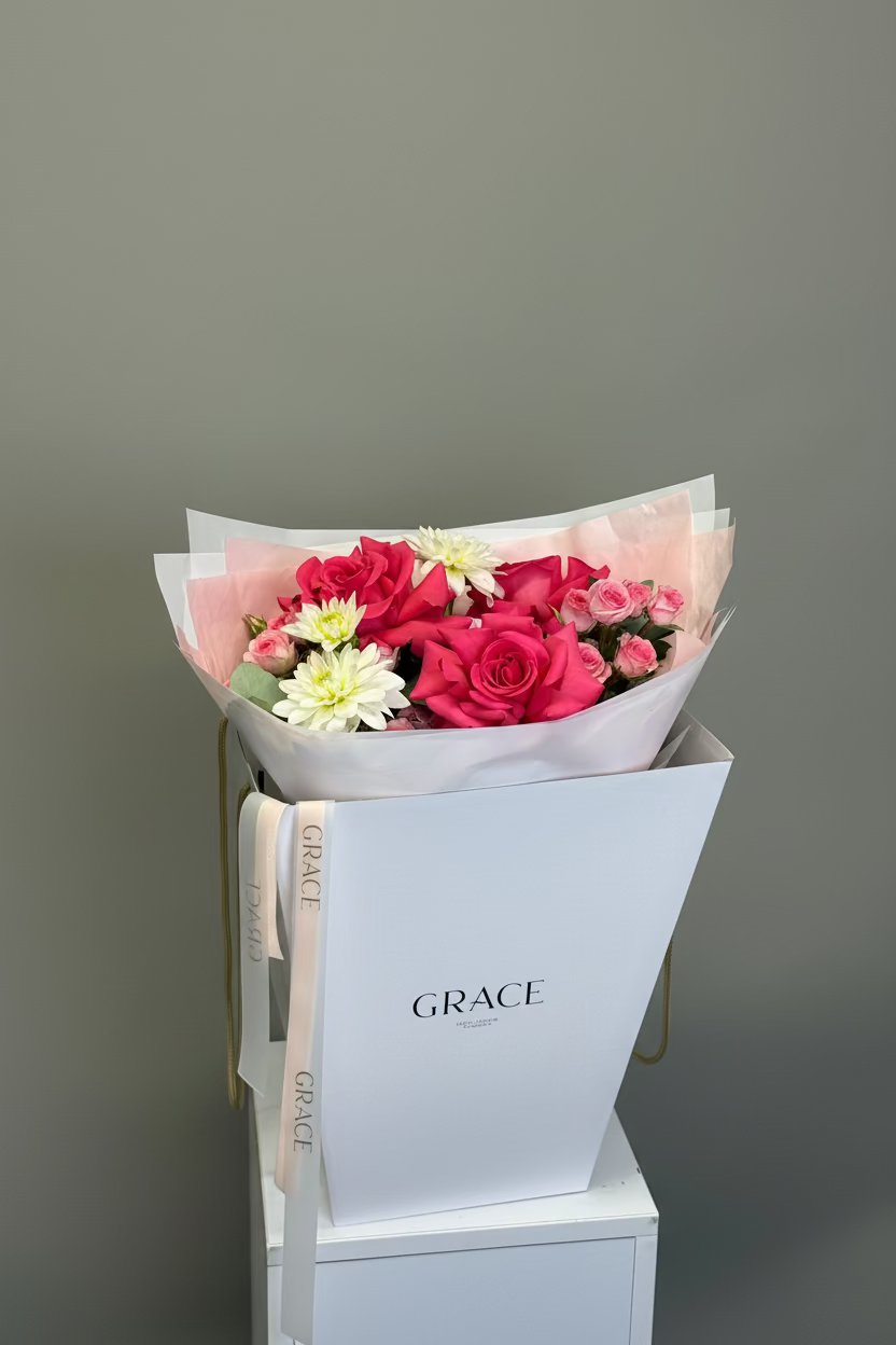 A floral bouquet with pink and red roses, white chrysanthemums, and green leaves, wrapped in pale pink tissue paper and presented in a white box branded GRACE. The box sits on a white pedestal against a plain gray background.
