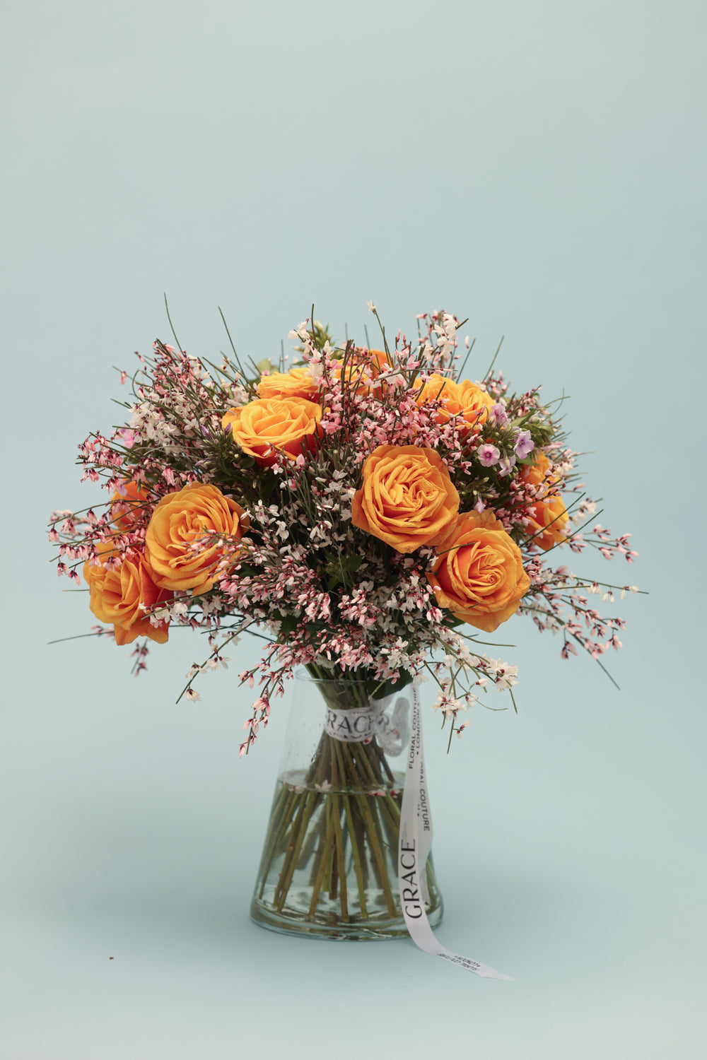 A vibrant bouquet of bright orange roses and delicate pink waxflower arranged in a clear glass vase, set against a light blue background.