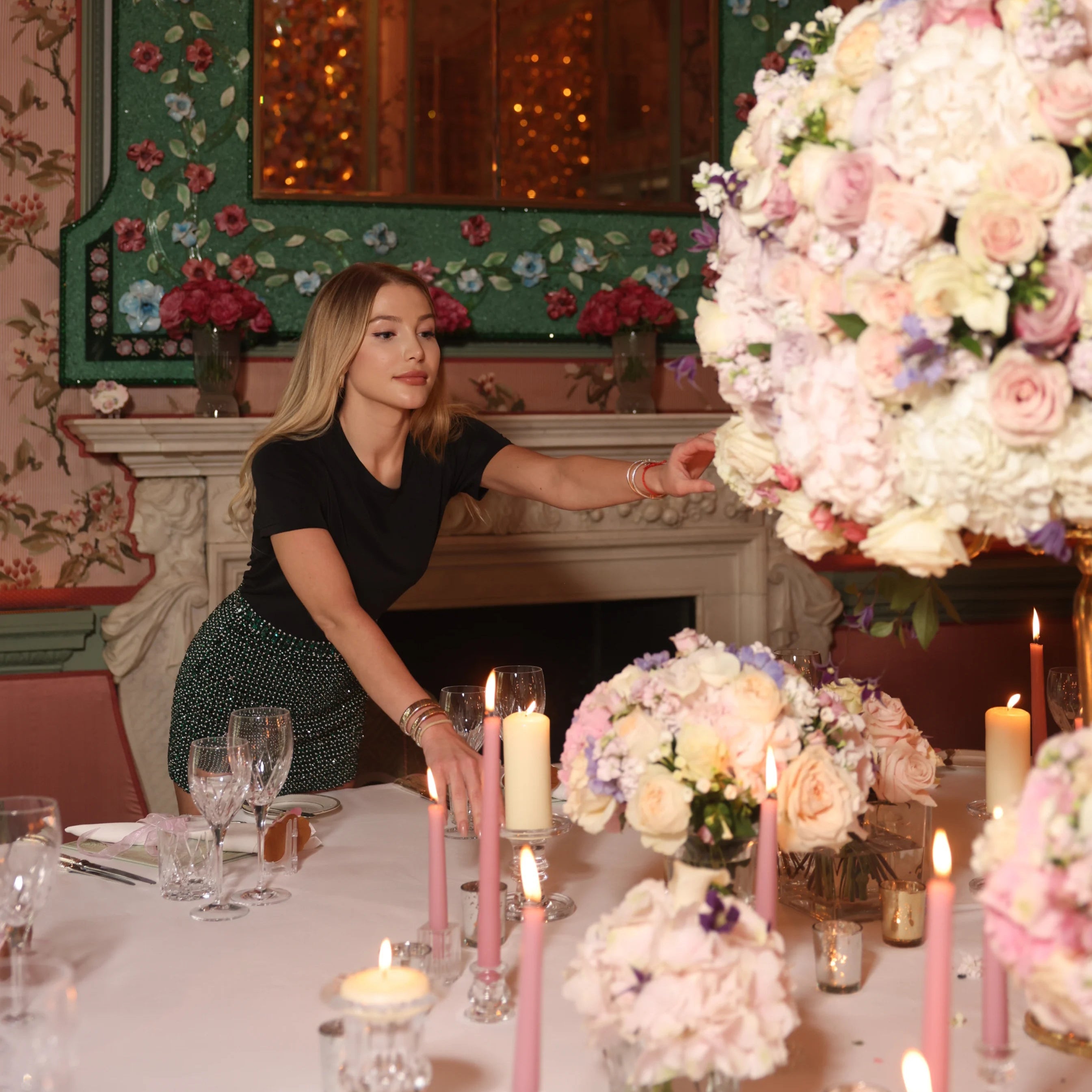 Woman arranging flowers at a decorated table with candles and floral arrangements.