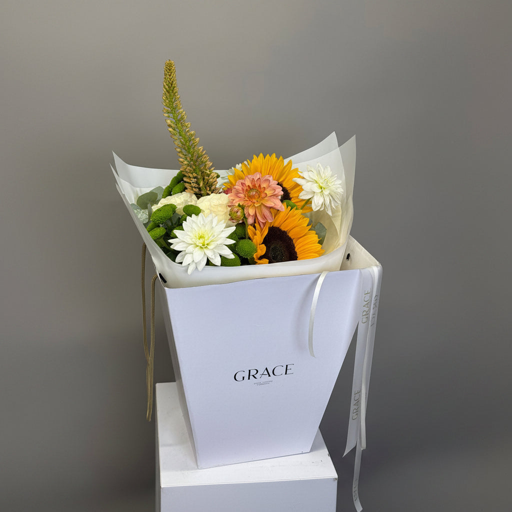 A floral arrangement in a white gift bag with the brand name GRACE printed on the front. The bouquet includes yellow sunflowers, white daisies, a peach/orange dahlia, green foliage, and a tall green ornamental grass, wrapped in white tissue paper and set against a neutral gray background.